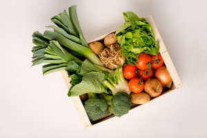 Assorted fresh vegetables including, tomatoes, fennel, potatoes, onions, broccoli, leeks, celeriac, and salad greens in a wooden crate over a white background.