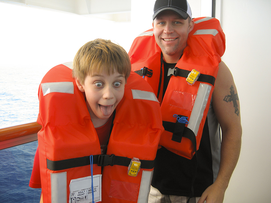 Father and his son put on life vests during cruise ship safety drill before departing. Ocean in background. Son makes silly face just as mom snaps the vacation photo.